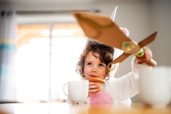 A portrait of happy small girl with a paper sword sitting at the table at home, eating muffins.