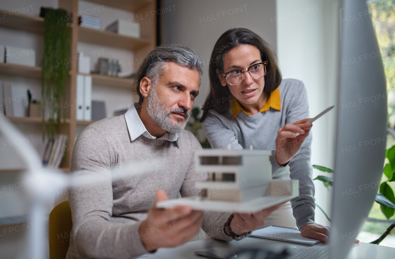 Mature architects with model of house sitting at the desk indoors in office, working.