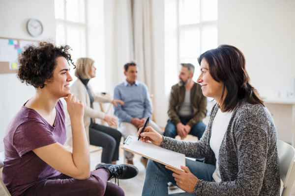 A senior counsellor with clipboard talking to a young woman during group therapy.