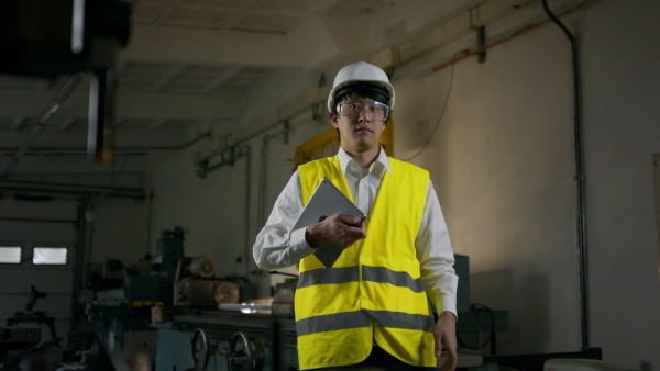 A young industrial Japanese man engineer with protective wear using tablet indoors in metal workshop.