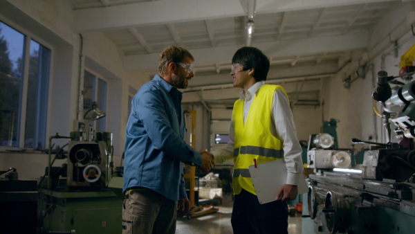 Japanese engineer shaking hands with a mature wroker indoors in metal workshop.