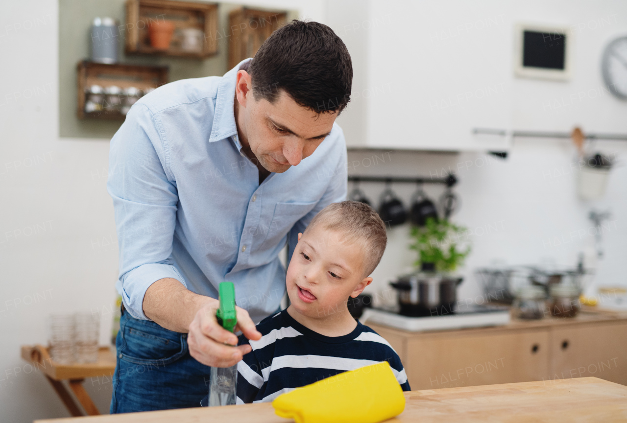 A father with happy down syndrome son indoors in kitchen, cleaning table.