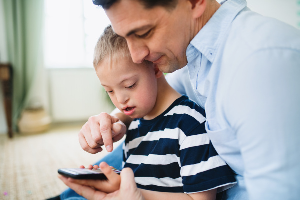 A father with happy down syndrome son indoors at home, using smartphone.