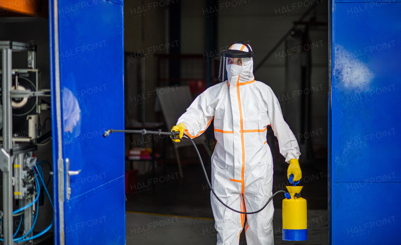 Front view portrait of man worker with protective mask and suit disinfecting industrial factory with spray gun.