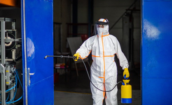 Front view portrait of man worker with protective mask and suit disinfecting industrial factory with spray gun.