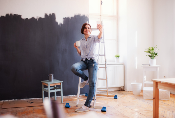 A portrait of young creative woman with a cup of coffee and smartphone painting wall black. A startup of small business.
