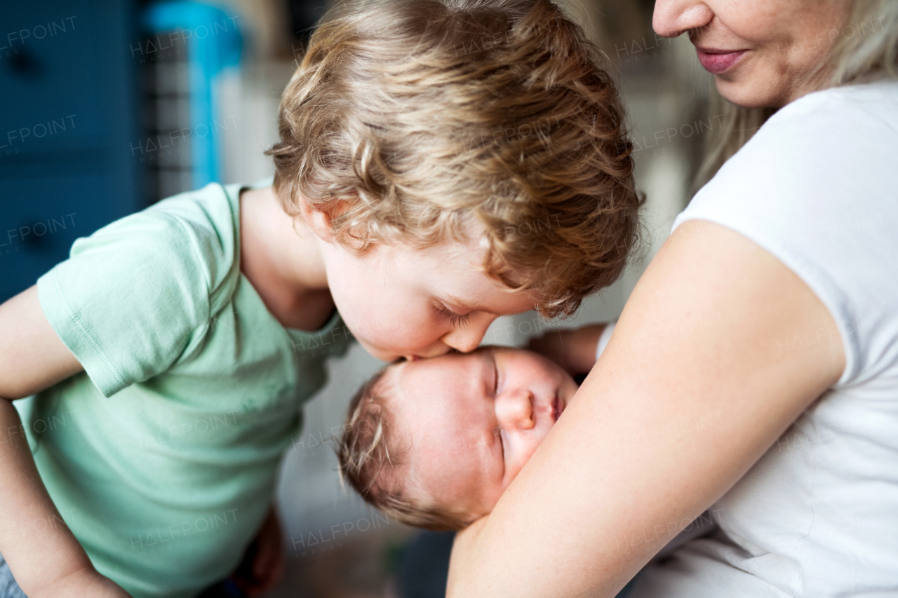 A small boy kissing a sleeping newborn baby brother at home, a mother holding him.