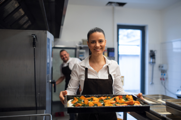 A happy female cook looking at camera and holding tray with baked pumpkin pieces in commercial kitchen.