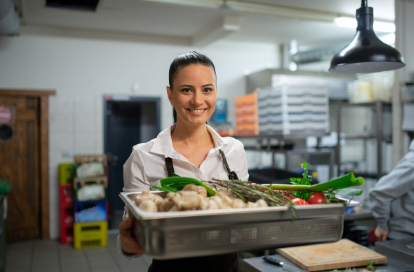 A female chef carrying tray with fresh vegetables and looking at camera in restaurant kitchen.