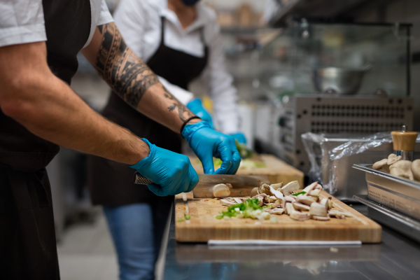 An unrecognizable chef with gloves cutting vegetables indoors in restaurant kitchen.