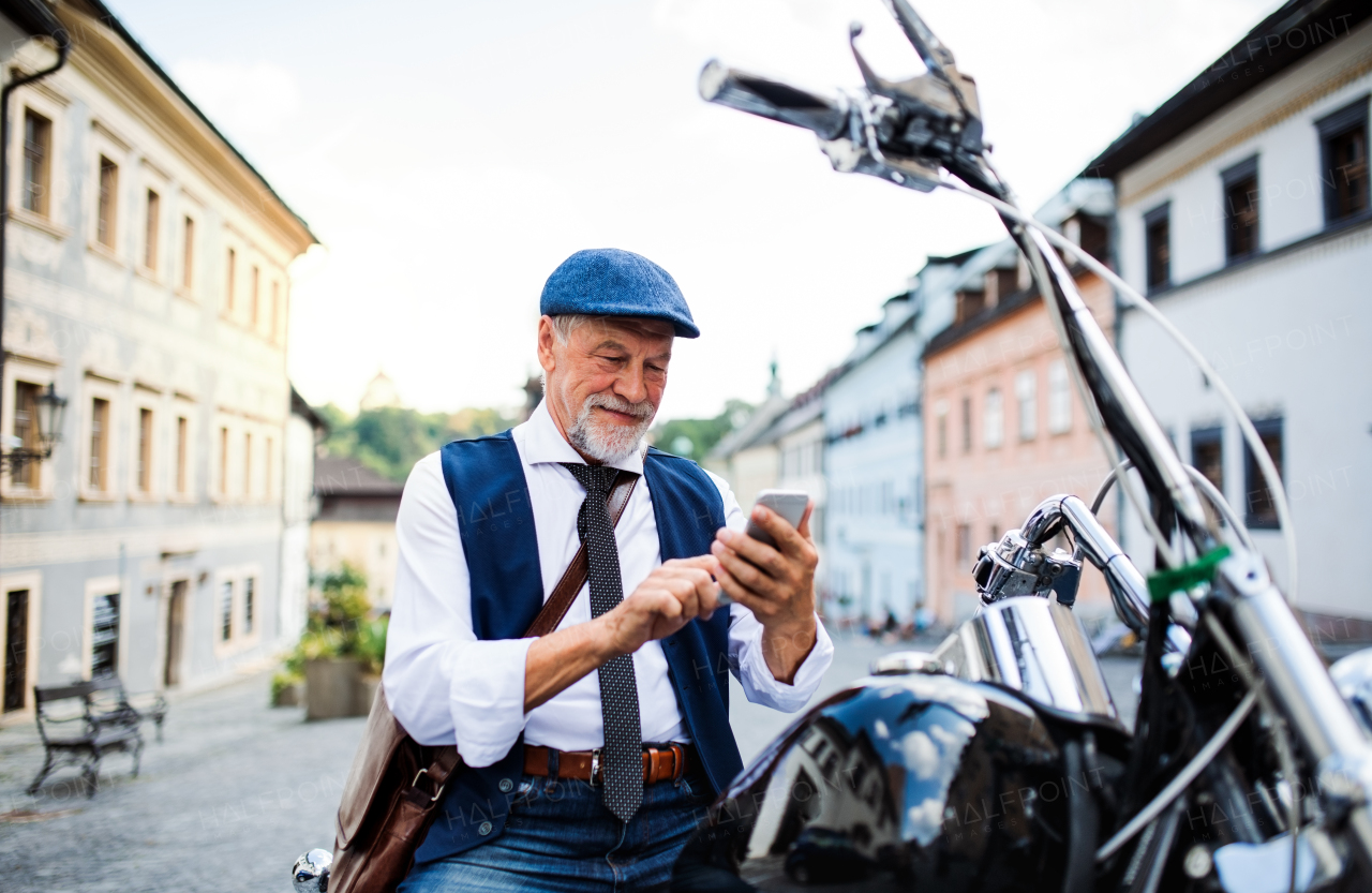 A cheerful senior businessman with motorbike in town, using smartphone.