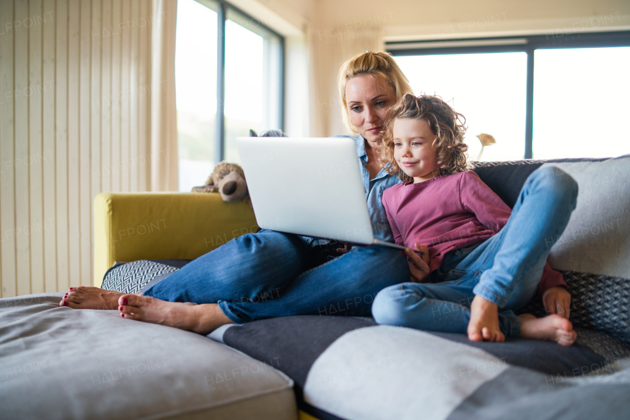 A cute small girl with mother sitting on sofa indoors at home, using laptop.