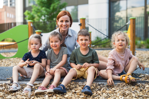 Group of small nursery school children with teacher sitting outdoors on playground, looking at camera.