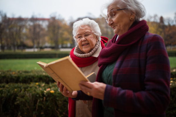 Happy senior women friends on a walk outdoors in town park in autumn, reading book and talking.