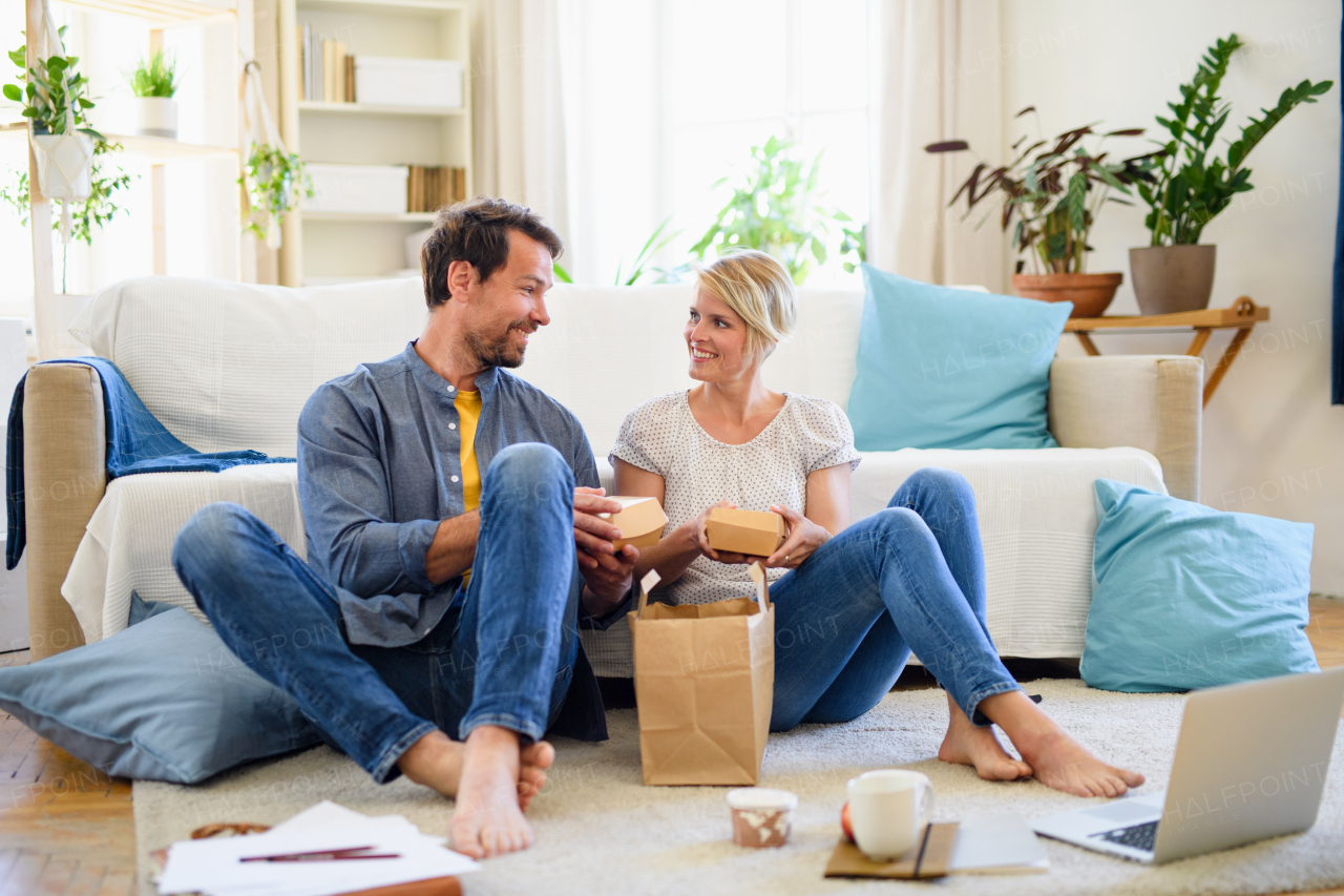 Front view of happy couple sitting on floor indoors at home, eating hamburgers.