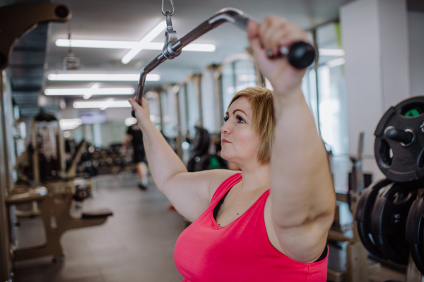 A mid adult plus size woman working out on a lat pulldown machine indoors in gym