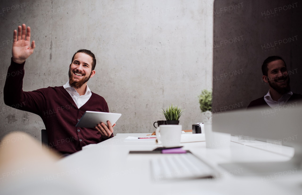 A young businessman with tablet sitting at the desk in office, greeting somebody.
