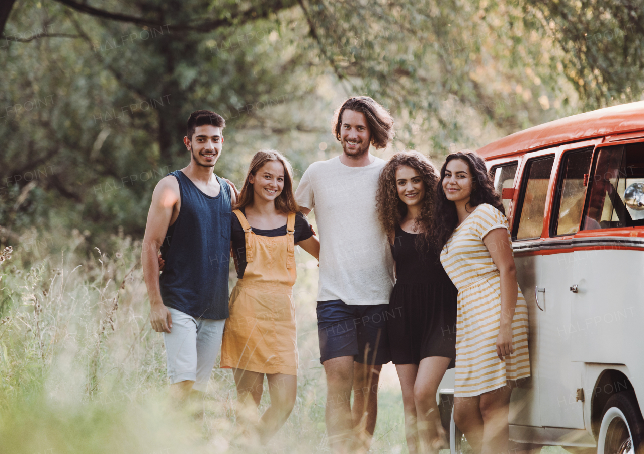 A group of young friends standing by a retro minivan on a roadtrip through countryside.