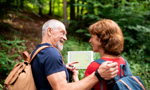 A senior tourist couple with backpacks on a walk in forest in nature, using map on tablet.