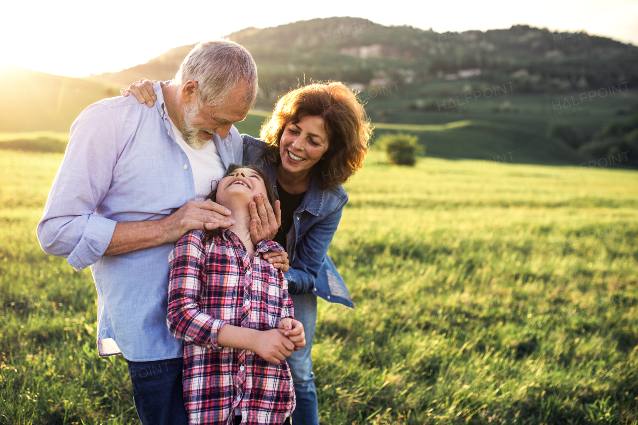 Happy senior couple with granddaughter standing outside in spring nature, looking at each other at sunset. Copy space.
