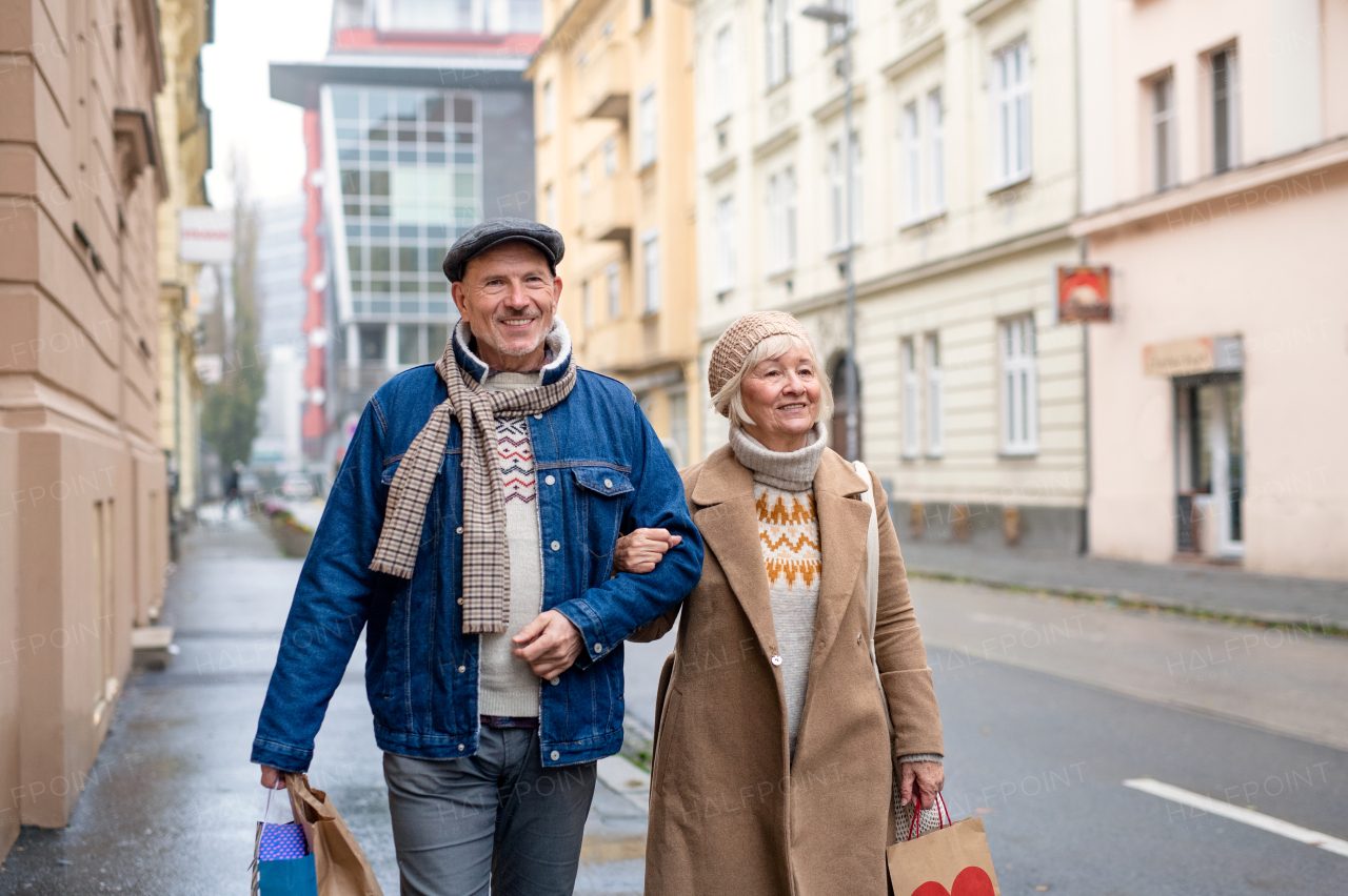 Portrait of happy senior couple walking outdoors on street in city, carrying shopping bags.