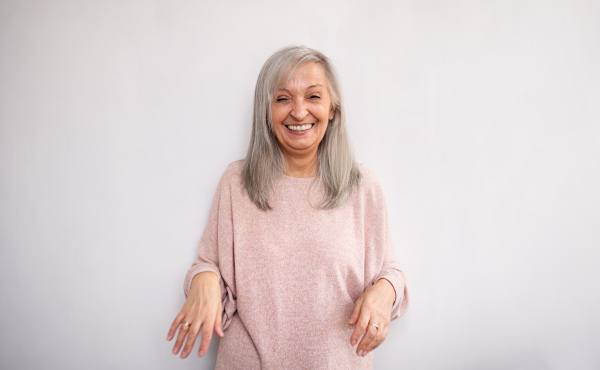 Front view portrait of senior woman standing indoors against light background, laughing.
