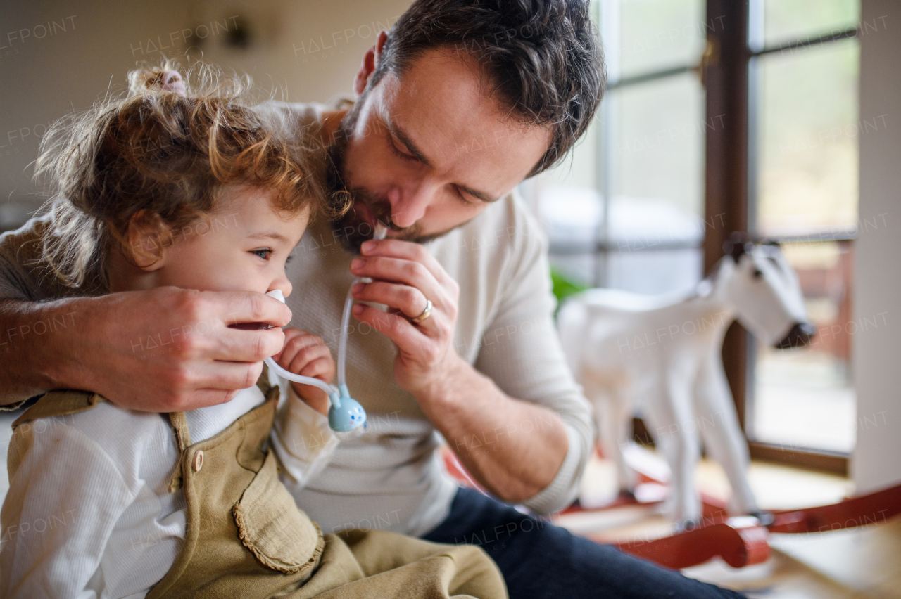 Mature father with small sick toddler daughter indoors at home, using nasal aspirator.