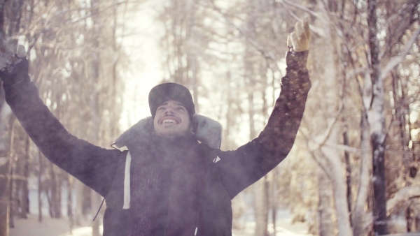 A portrait of young man standing outdoors in snow in winter forest, throwing snow. Slow motion.