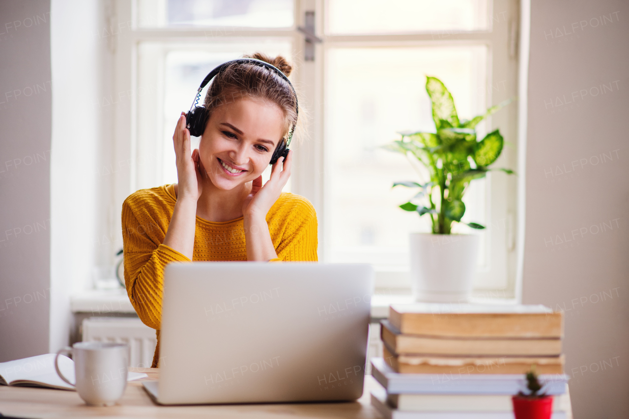 A young happy college female student sitting at the table at home, using laptop and headphones when studying.