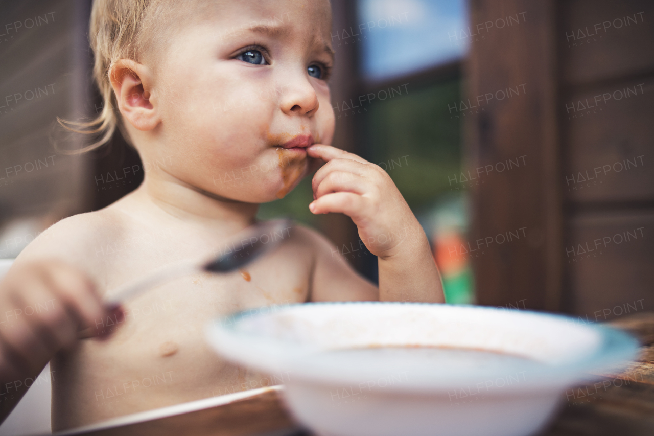 Cute topless small girl standing by a table on a patio in summer, eating soup.