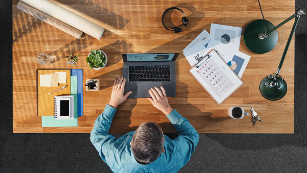 A top view of businessman working on laptop computer at desk with paperwork in home office.