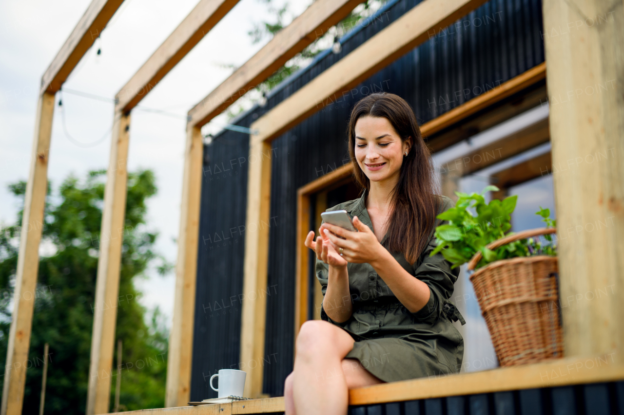 Happy young woman with smartphone outdoors, weekend away in container house in countryside.