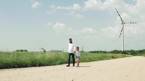 A mature father with small daughter on field on wind farm, walking.