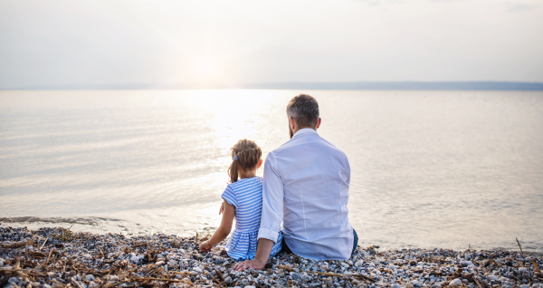 Rear view of father and small daughter on a holiday sitting by the lake or sea.