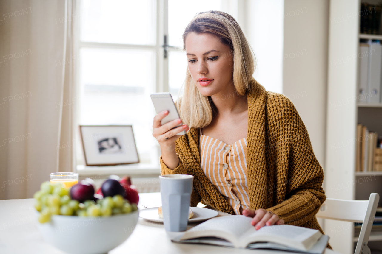 Young woman sitting at the table indoors at home, using smartphone when relaxing.