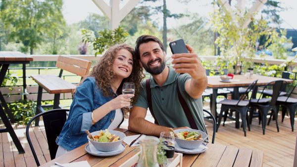 Happy couple sitting outdoors on terrace restaurant, taking selfie with smartphone.