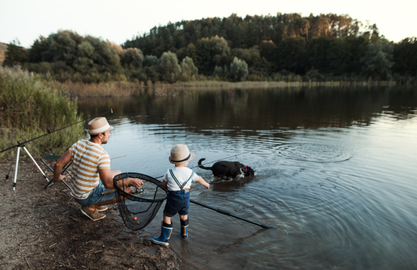 A mature father with a small toddler son and a dog outdoors fishing by a river or a lake.
