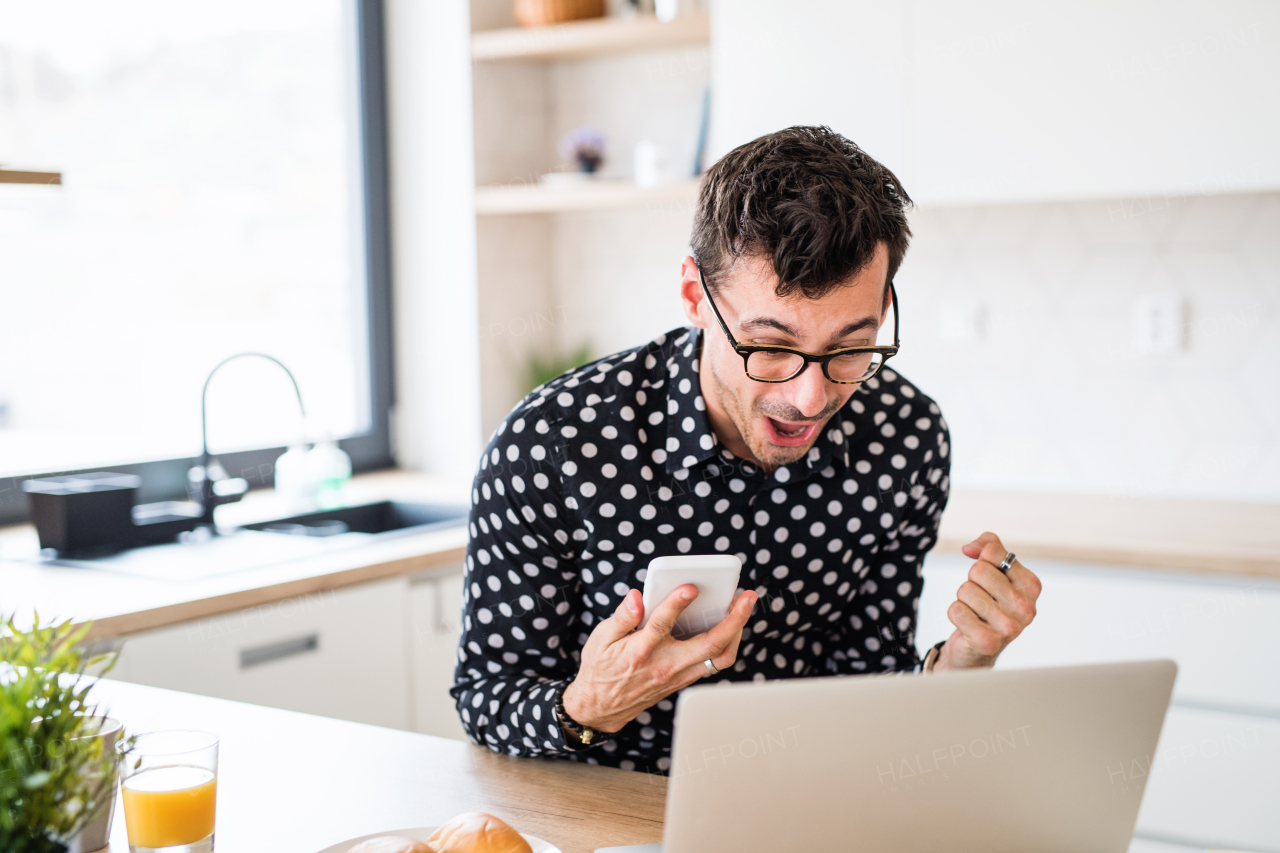 Young man with laptop and smartphone sitting in kitchen, expressing excitement. A home office concept.