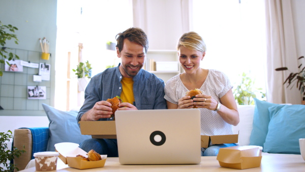 Front view of happy couple with laptop sitting on sofa indoors at home, eating hamburgers.