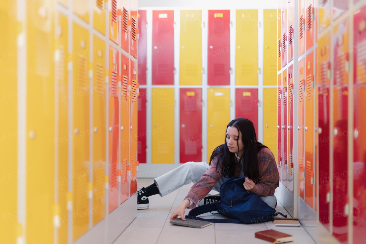 A teenage student sitting in corridor near colorful lockers adn packing book to backpack in campus hallway, back to school concept.