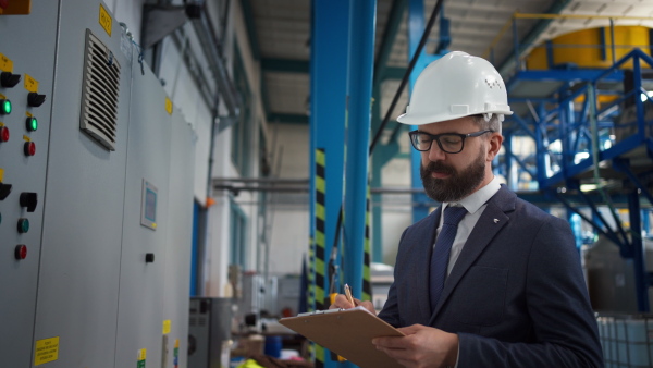 A chief Engineer in the hard hat walks through industrial factory while holding tablet.