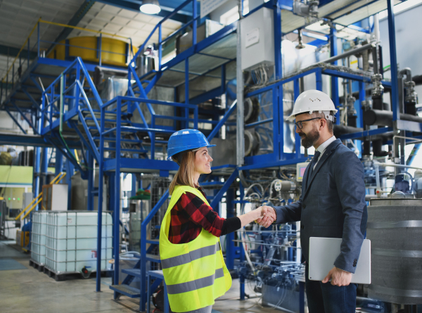 An engineer and industrial worker in uniform shaking hands in large metal factory hall and talking.