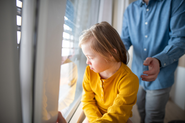 A sad little girl with Down syndrome looking throug window with her father at background.