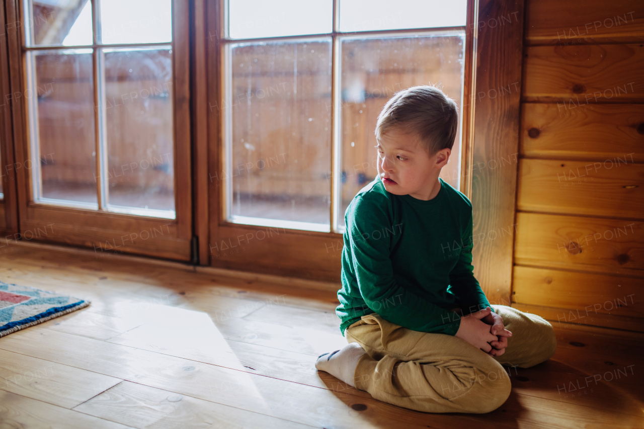 A sad little boy with Down syndrome sitting on floor at home.