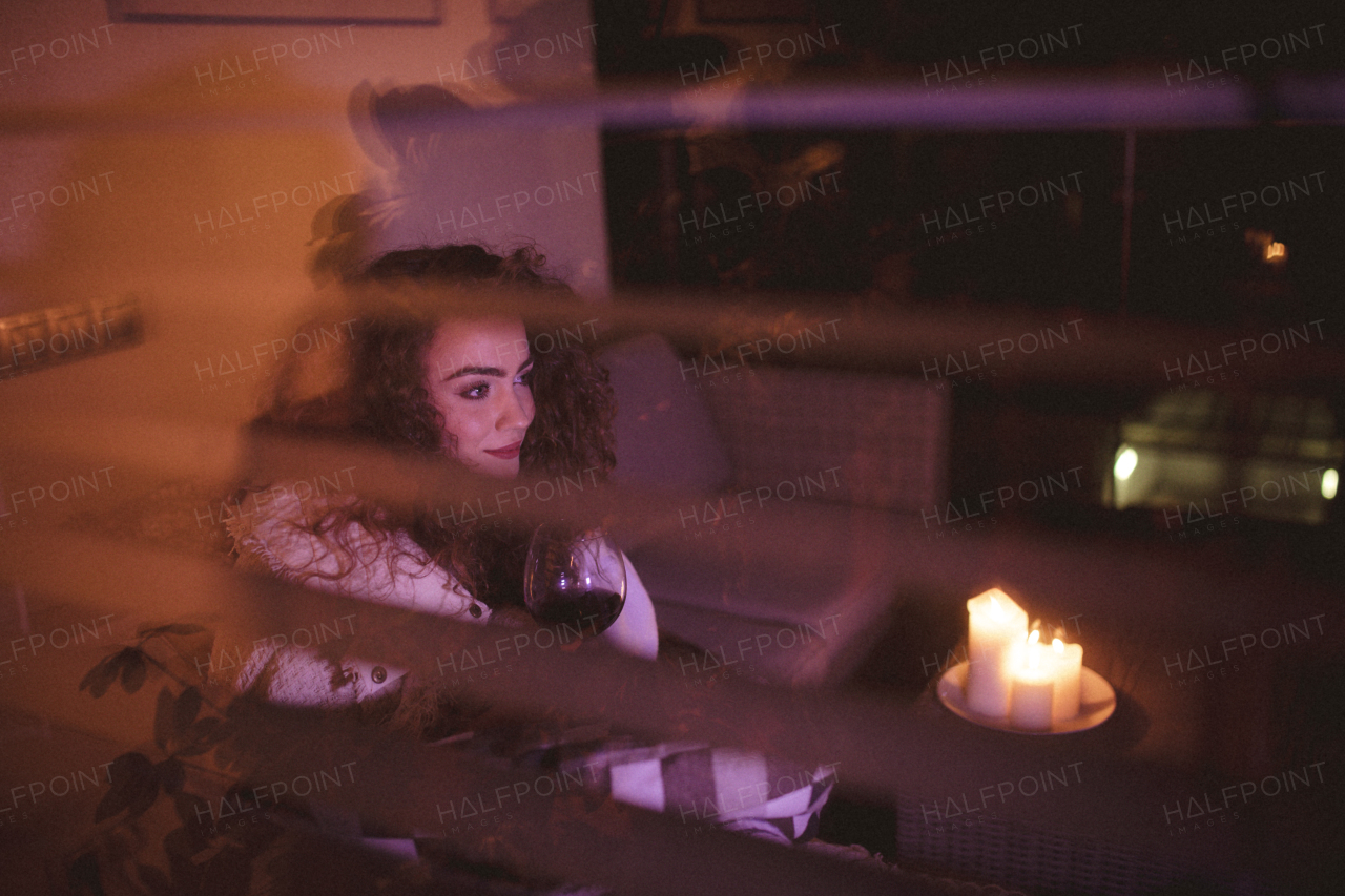 A happy young woman wrapped in blanket sitting on terrace and drinking wine in the evening shot through window.