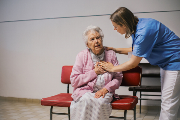 Senior patient with heart attack feeling chest pain, sitting in front of emergency room. Nurse helping elderly woman in hospital gown. Panic attack.