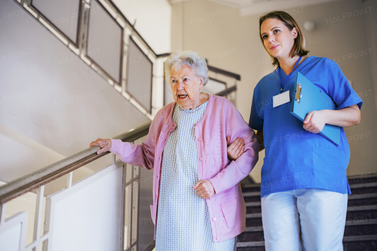 Nurse helping senior patient walk down the stairs. Hospitalized elderly woman in hospital gown is anxious. Emotional support from healthcare workers.