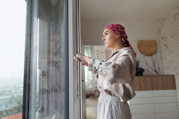 Young unhappy woman with cancer standing in front of the window.