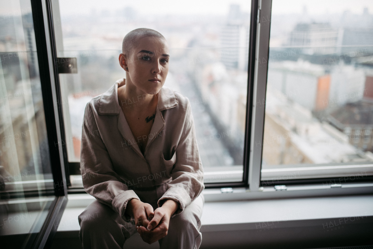 Young unhappy woman with cancer sitting in window and looking at view.