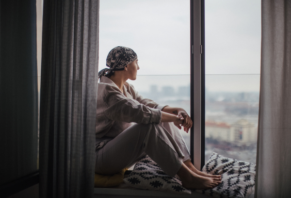 Young unhappy woman with cancer sitting in window and looking at view.